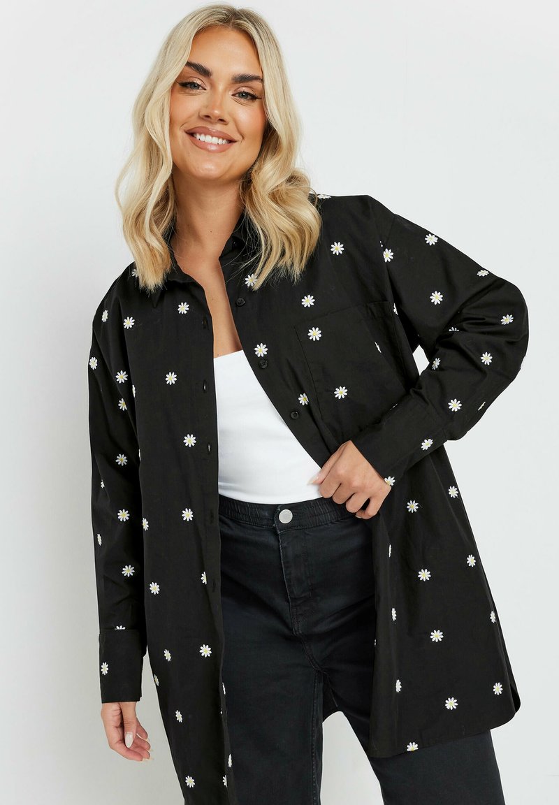 Camicia nera con bottoni, caratterizzata da ricami bianchi a forma di margherita, maniche lunghe e una vestibilità comoda. Indossata sopra un top bianco e pantaloni neri.