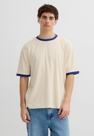 Joven con cabello rizado castaño, vistiendo una camiseta beige con cuello y bordes de las mangas azules, combinada con jeans azul claro, sobre un fondo liso.