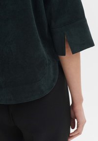 Dark green corduroy top with 3/4 sleeves featuring side slits and a rounded hem. Texture is ribbed, providing a soft appearance.