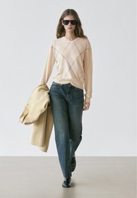 Vrouw loopt en draagt een beige argyle trui, donkerblauwe spijkerbroek, zwarte loafers, zwarte zonnebril, houdt een beige jas in haar linkerhand tegen een effen achtergrond.