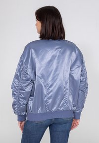 Jasnoniebieska satynowa kurtka bomber z ściągaczami na mankietach, w oversize'owym kroju, z marszczeniem na rękawach. Nosi ją osoba w niebieskich dżinsach, widok z tyłu.