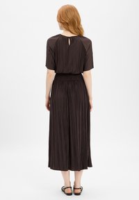 Plisserad mörkbrun klänning med nyckelhålsrygg, korta ärmar och en rynkad midja. Mjukt tyg som faller till mitten av vaden. Svarta flata sandaler.