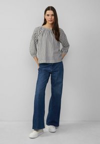 Gestreepte, lichte top met driekwartmouwen, gecombineerd met wijde blauwe jeans en witte sneakers tegen een grijze achtergrond.