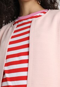 Ljusrosa texturerad kofta över en röd-och-vit randig T-shirt med rosa krage. Designen är enkel och samtida.