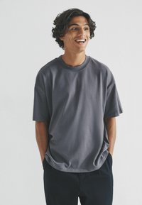 Graues Baumwoll-T-Shirt mit Rundhalsausschnitt, abgesenkten Schultern und lässiger Passform. Getragen mit dunklen Hosen, vor einem schlichten Hintergrund präsentiert.