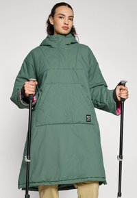 Parka acolchada verde con un gran bolsillo frontal, un cuello alto y acentos rosas en el interior de las mangas. Incluye bastones de trekking.