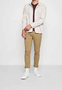 Ljusbeige jacka, vinröd skjorta, khaki-byxor och vita sneakers. Outfiten har en avslappnad design med knappar och uppvikta ärmar.