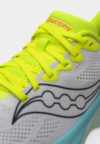 Λευκό και νέον κίτρινο αθλητικό παπούτσι Saucony με πλεκτό επάνω μέρος, φωτεινά κορδόνια και μπλε ενδιάμεση σόλα, κοντινή λήψη στο πλάι και στο λογότυπο της γλώσσας.