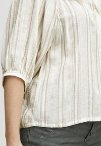 Blusa ligera de crema con rayas verticales en beige, cierre de botones en la parte delantera y mangas ligeramente abullonadas. Tejido texturizado con un patrón tejido.