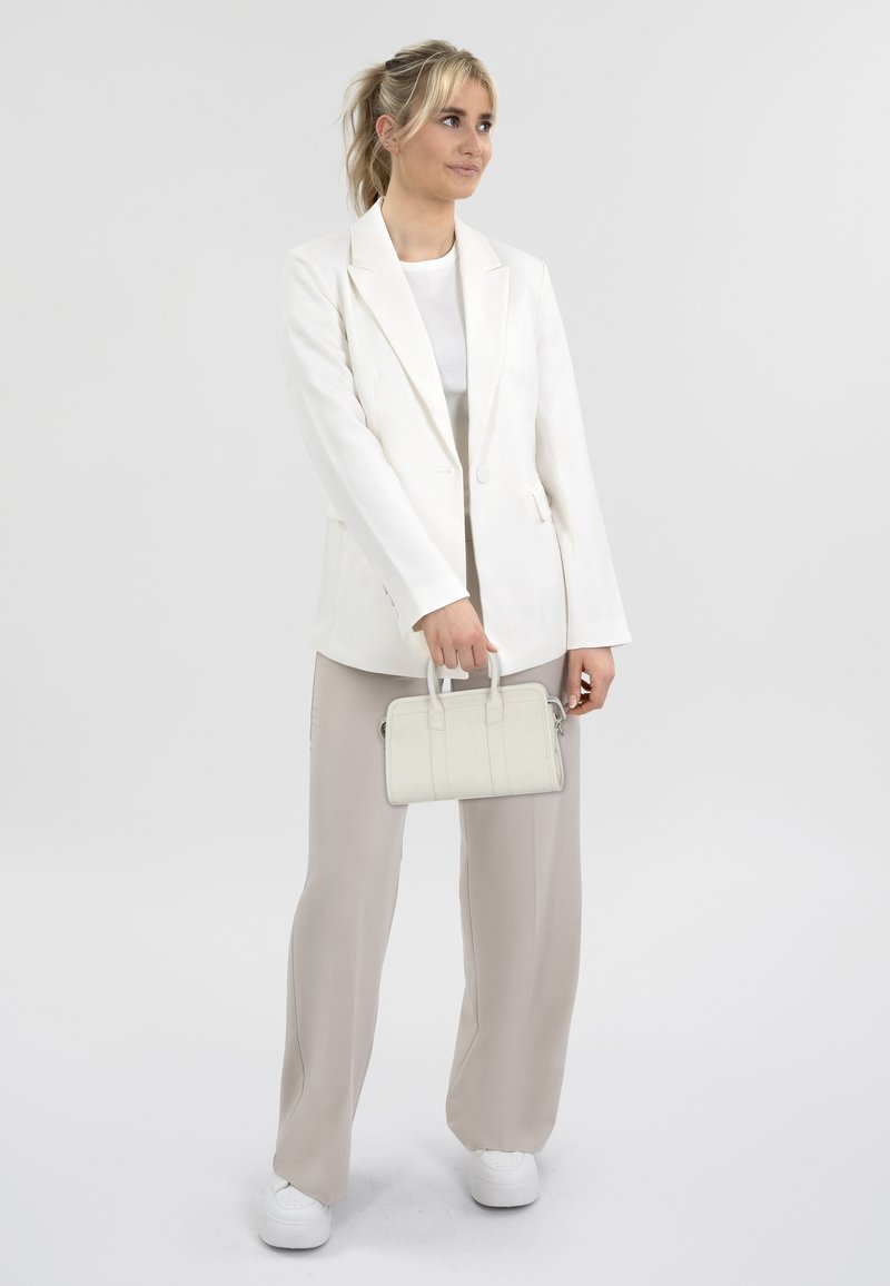 Blazer bianco su un top grigio chiaro, abbinato a pantaloni beige e sneakers bianche; tiene in mano una piccola borsa bianca.