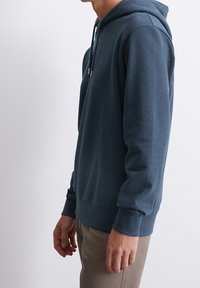 Mörkblå hoodie i mjukt material, med en dragsko i huvan, ribbade muddar och rak nederkant. Minimal branding på den nedre sidan.