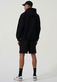 Svart oversized hettegenser med hette, kombinert med svarte shorts. Hvite og grå sneakers med teksturert såle og svarte sokker med tekst.