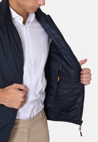 Gilet imbottito blu navy con interno trapuntato, dotato di una tasca laterale con zipper arancione, tessuto leggero e chiusura frontale con zip.