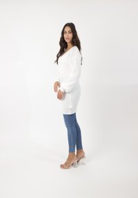 Witte gebreide oversized trui met lange mouwen, gecombineerd met blauwe skinny jeans en doorzichtige sandalen met hakken, tegen een witte achtergrond.