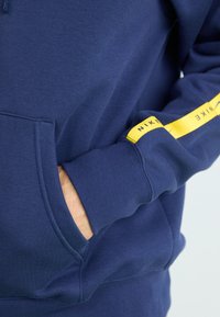 Navy blue hoodie with a front pocket, ribbed cuffs, and a yellow Nike logo stripe running down the right sleeve. Soft texture.