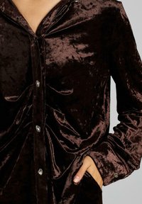 Camicia in velluto marrone con chiusura a bottoni, maniche lunghe e tasca. Presenta bottoni argentati lucidi e leggere variazioni di texture.