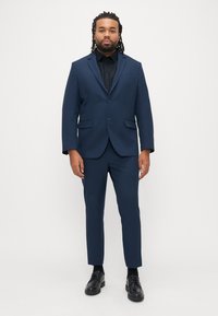 Marineblauer Anzug mit tailliertem Schnitt, Ein-Knopf-Verschluss und gekerbtem Revers. Schwarzes Hemd darunter, kombiniert mit glänzend schwarzen Schuhen.