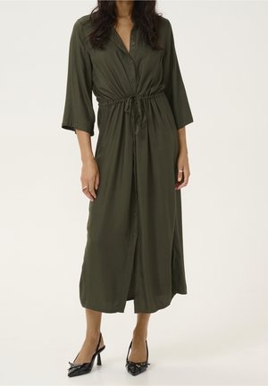 Femme portant une robe midi vert olive avec boutonnage sur le devant, taille à cordon, manches 3/4, et des escarpins slingback noirs à bout pointu.