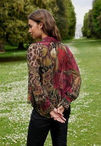 Giacca trasparente con motivo floreale nei toni di rosso e verde, dotata di colletto rotondo e maniche plissettate, abbinata a pantaloni neri aderenti.