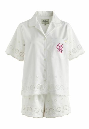 Femme debout pieds nus sur la rambarde du balcon, portant une chemise blanche à manches courtes en broderie anglaise et un short assorti, avec des collines et des bâtiments en arrière-plan.