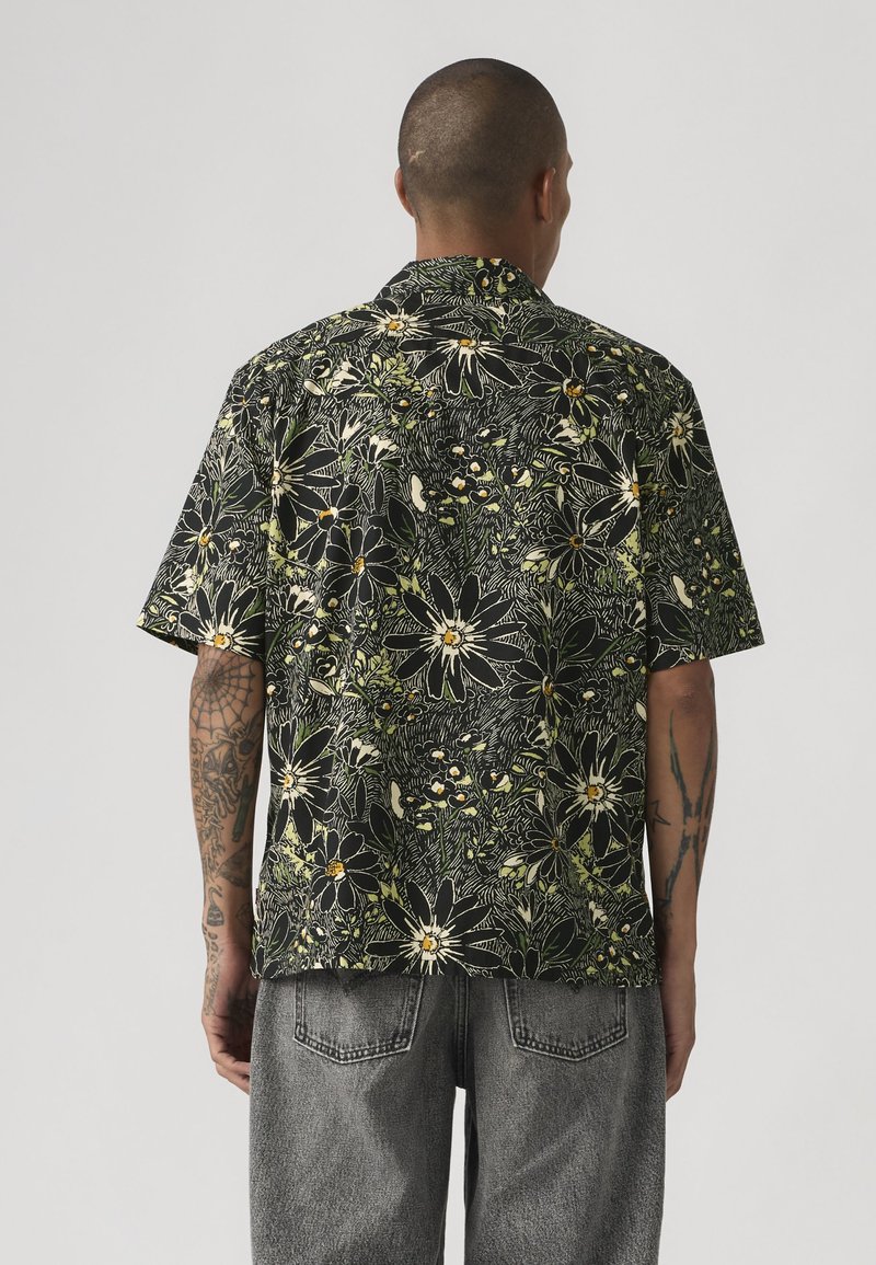 Uomo con capelli corti e tatuaggi sulle braccia, che indossa una maglietta nera a maniche corte con una grande stampa floreale bianca e gialla e jeans in denim grigi.