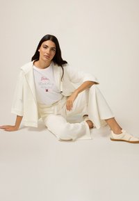 Tenue blanche composée d'une veste ample avec poches et d'un pantalon à jambe large assorti. En dessous, un t-shirt graphique avec du texte rouge et argent.