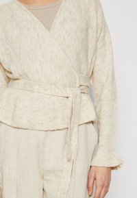 Beige wrap cardigan in soft, textured fabric. Features a tied waist, ribbed hem, and flared cuffs. Paired with matching light-colored trousers.