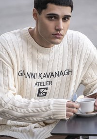 Cremfarbener Kabelstrickpullover mit gesticktem Text "GIANNI KAVANAGH ATELIER." Modell hält eine weiße Tasse auf einem schwarzen Tisch, strukturierter Stoff sichtbar.
