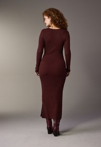 Geripptes kastanienbraunes Kleid mit langen Ärmeln, figurbetontem Schnitt und seitlichen Schlitzen, kombiniert mit kniehohen burgunderroten Stiefeln, vor neutralem Hintergrund.