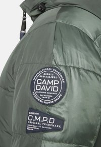 Grön puffjacka med två runda patchar: en med "CAMP DAVID" och en annan med "C.M.P.D." i kontrasterande svart och vit text.