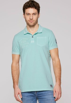 Uomo che indossa una polo azzurro chiaro con taschino sul petto e jeans, in piedi davanti a uno sfondo grigio chiaro uniforme.