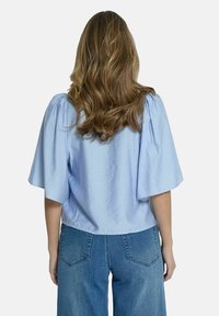 Blouse bleu clair à manches courtes avec épaules froncées, ourlet fini, portée avec un jean en denim bleu taille haute, vue de dos.