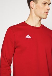 Sweatshirt rouge à manches longues en tissu doux, avec un col rond et un logo Adidas blanc sur la poitrine gauche.