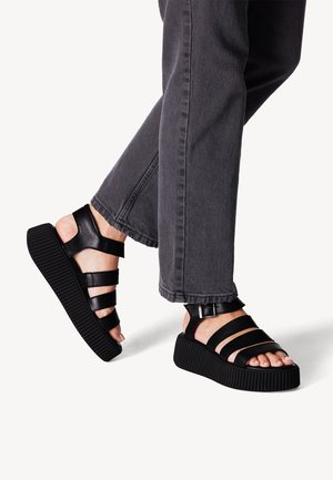 Sandali platform neri con molte cinghie larghe, realizzati in pelle liscia, con suola rigata, indossati con pantaloni in denim grigi.