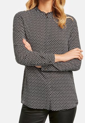Femme portant un chemisier à manches longues noir et blanc à motifs avec un petit col à volants et un pantalon noir, les bras croisés sur la poitrine.