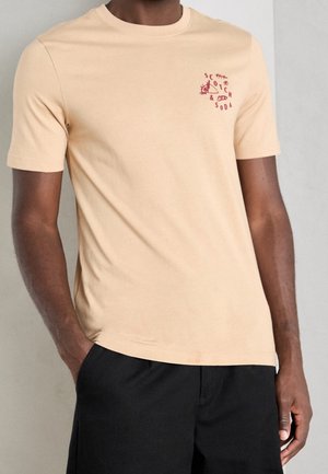 Mężczyzna ubrany w beżowy T-shirt z krótkim rękawem z małym czerwonym logo "Scotch & Soda" na lewej piersi oraz czarne szorty, ręce w kieszeniach.