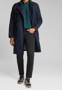 Abrigo tipo gabardina azul marino sobre una camisa verde abotonada, jeans negros y zapatillas grises. Cuenta con botones cruzados y mangas con cinturón.