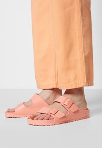 Chanclas de goma de color melocotón con diseño de punta abierta, correa ajustable con hebilla y suela texturizada para agarre. Llevadas con pantalones naranja claro.