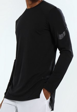 Mann trägt ein schwarzes, langärmeliges Sportshirt mit reflektierendem Logo auf dem linken Ärmel, kombiniert mit weißer Hose.