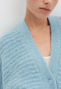 Cardigan fuzzy azzurro chiaro con un motivo testurizzato, scollo ampio e chiusura con un singolo bottone rotondo. Tessuto morbido e lavorato a maglia visibile.