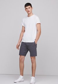 T-shirt blanc à col rond associé à un short gris foncé. Le mannequin se tient de manière décontractée, les mains dans les poches, portant des baskets montantes blanches.