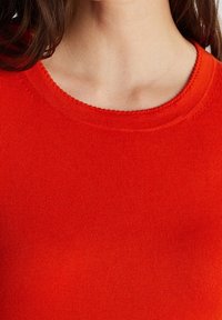Red knit sweater with a round neckline, featuring fine ribbed texture and smooth fabric. The color is vibrant and solid without patterns.