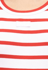 Κοντινή όψη λευκού ριμπ μπλουζιού με οριζόντιες κόκκινες ρίγες και στρογγυλή λαιμόκοψη, με μικρό λευκό σήμα στο στήθος.