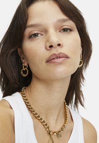 Collana in oro con grandi maglie, finitura lucida, abbinata a orecchini a cerchio in oro dello stesso stile. Il modello indossa una maglietta senza maniche bianca.