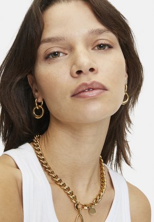 Collier en chaîne dorée avec de grands maillons, finition polie, associé à des boucles d'oreilles créoles dorées assorties. Le modèle porte un haut sans manches blanc.
