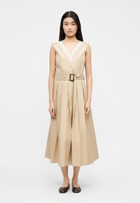 Donna che indossa un vestito midi beige senza maniche, plissettato, con una cintura larga e ballerine marroni, in piedi contro uno sfondo bianco neutro.