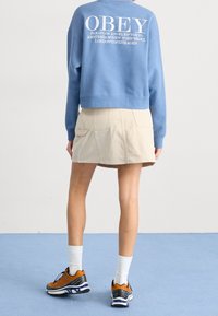 Ljusblå cropped sweatshirt med "OBEY" logotyp på baksidan, tillsammans med en beige denim minikjol och sneakers med orange detaljer på vita strumpor.