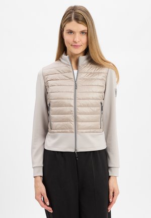Beige gewatteerde vest met hoge kraag, rits aan de voorkant en zijzakken, gecombineerd met een grijs longsleeve shirt en zwarte broek.