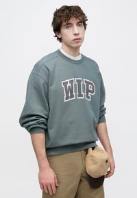 Γκρι φούτερ με την επιγραφή «WIP» σε σκούρα γράμματα, διαθέτει στρογγυλή λαιμόκοψη και φαρδιά μανίκια. Μπεζ καπέλο κρέμεται από τη ζώνη.