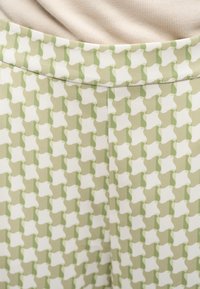 Κοντινό πλάνο υφάσματος με πράσινο και λευκό μοτίβο χάουντσουθ, με καμπύλη ραφή και μπεζ υπόβαθρο υφάσματος ορατό από πάνω.